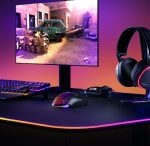 Gaming desk setup with RGB-lit edge, monitor showing an in-game action scene, colorful mechanical keyboard, and glowing mouse with a headset nearby.