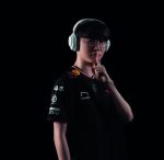 Esports player in a black jersey with sponsor logos wearing white over-ear headphones, shushing with a finger to the lips against a dark background.