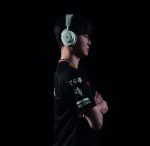 Side view of a young man wearing silver over-ear headphones, in a black esports jersey with logos, arms crossed against a dark background.