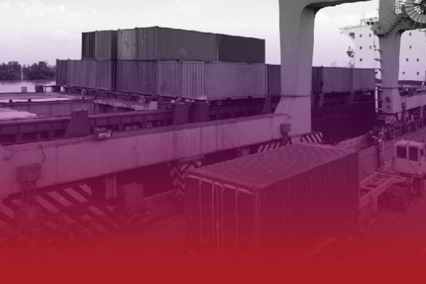 Stacks of shipping containers on a cargo terminal with a large gantry crane in the background.