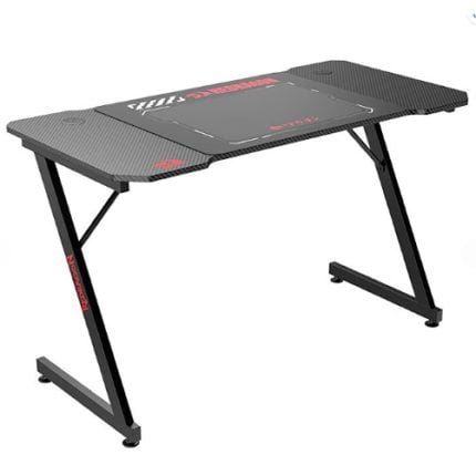 Black gaming desk with carbon-fiber textured top and angled twin-leg frame, shown from a side angle.