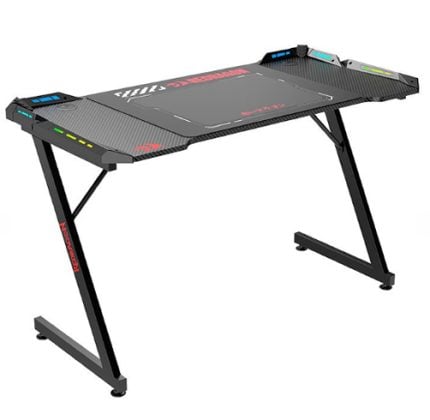 Black carbon-fiber-style gaming desk with angled X-frame legs and a wide top, featuring control panels along the edges for settings and lighting.