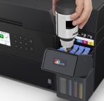 Person refilling the printer's ink tanks on an EcoTank model; front panel open showing cyan, magenta, yellow ink reservoirs and a refill bottle being squeezed into the tank.